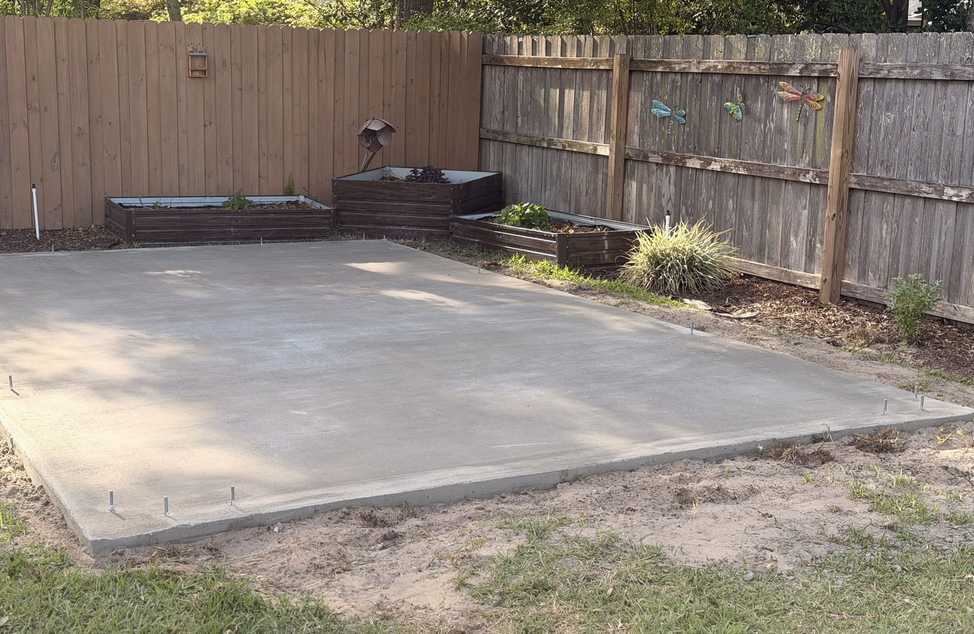 Concrete patio slab in a fenced backyard with low retaining blocks and patchy grass.