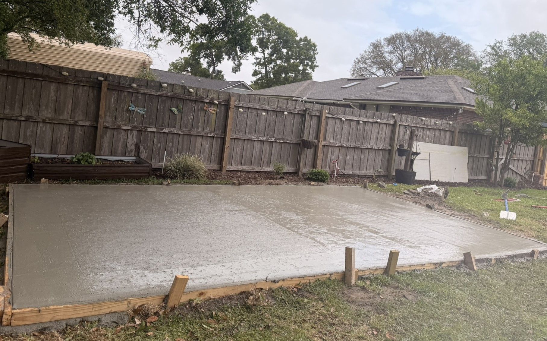 Wet concrete slab in a backyard beside a wooden fence and house, with trees in the background