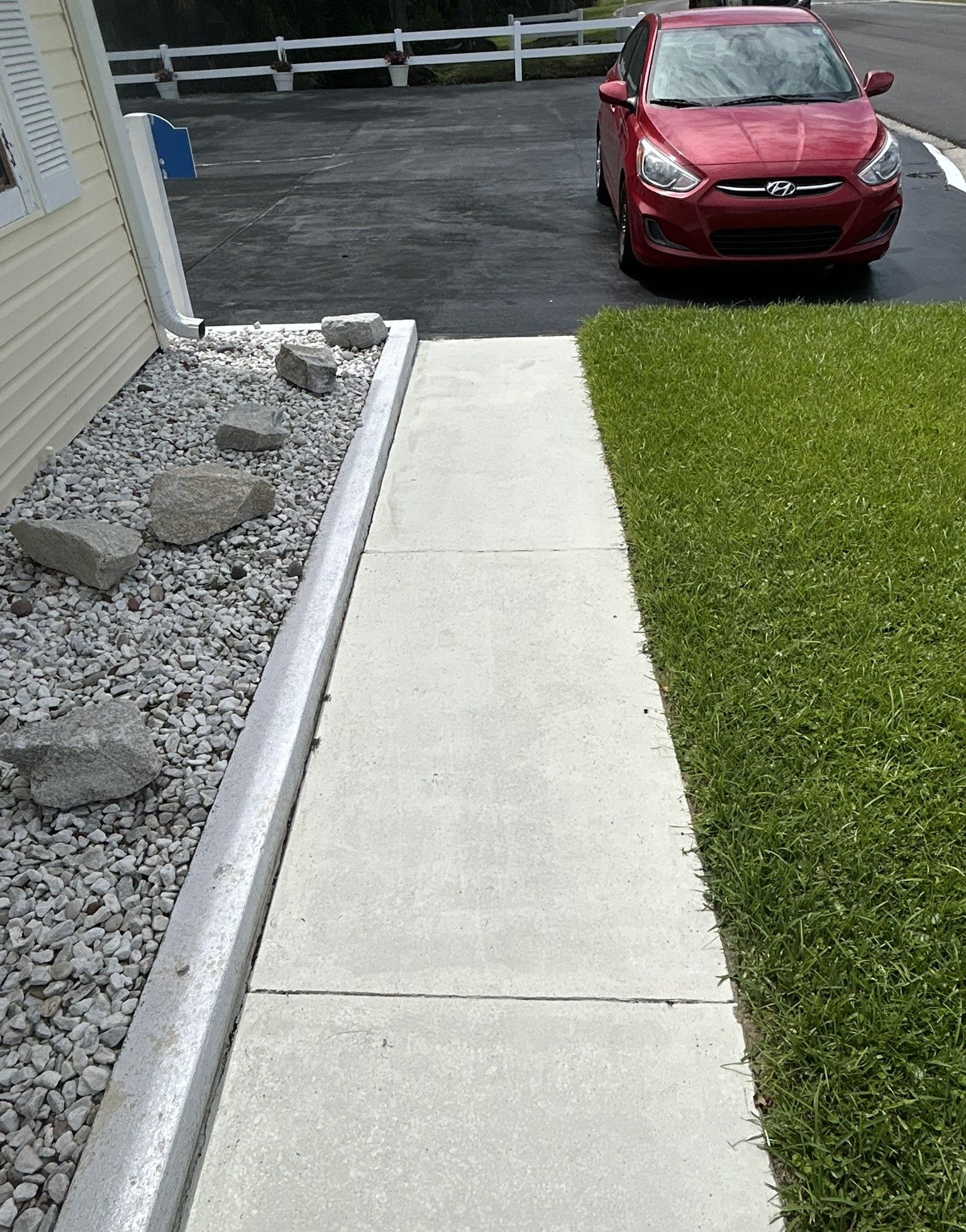 Concrete walkway beside lawn and gravel bed leading to a driveway with a red car parked ahead.