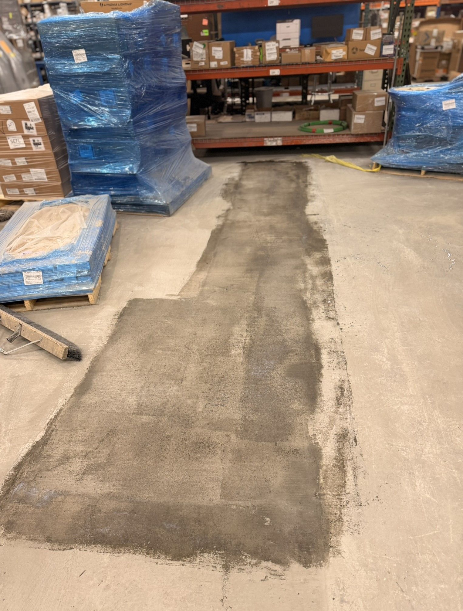 Warehouse aisle with a repaired concrete floor patch between stacked pallets and shelving.