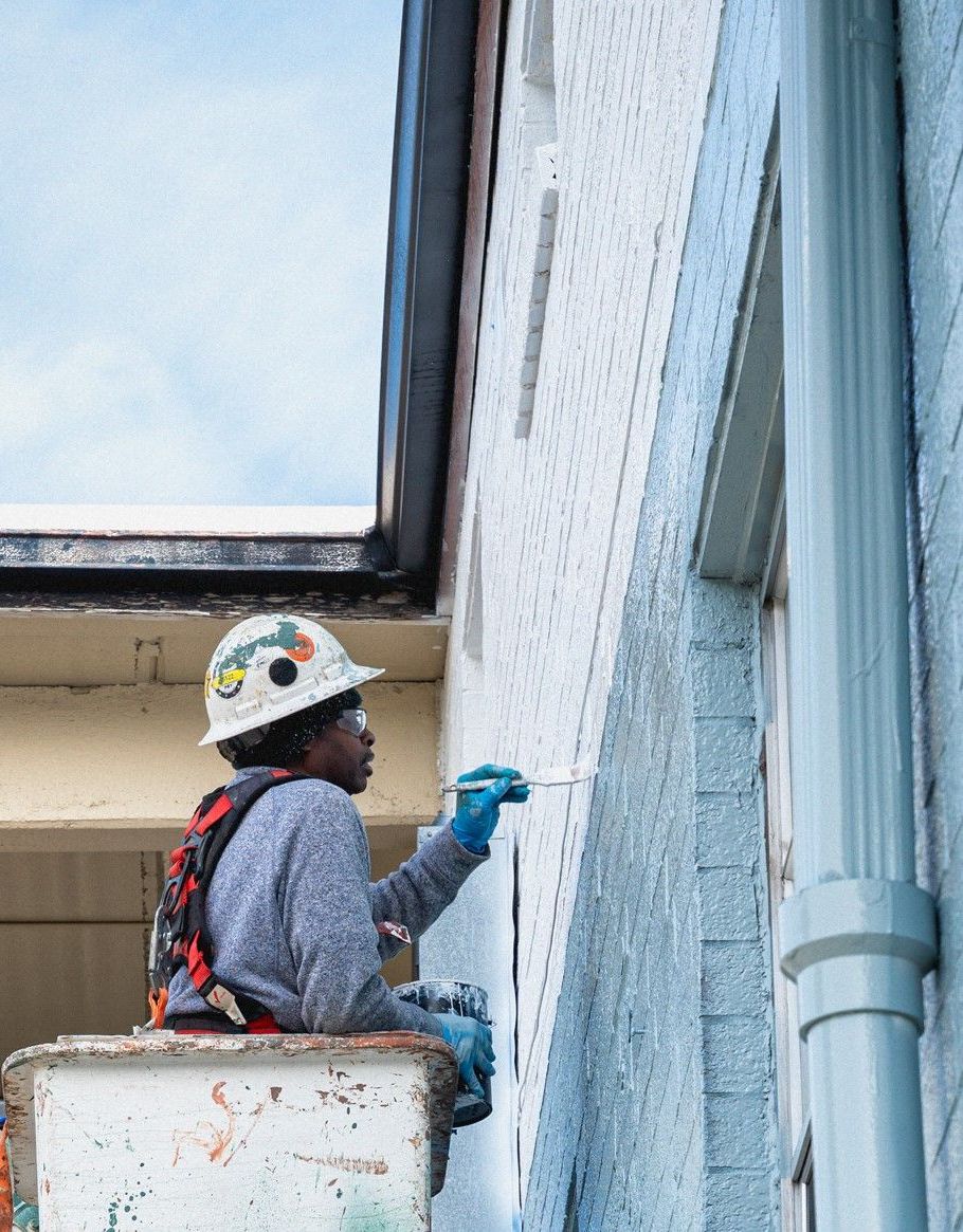 Construction worker paints a building's exterior while seated in a lift. He wears a hard hat and gloves, working on light-colored walls.