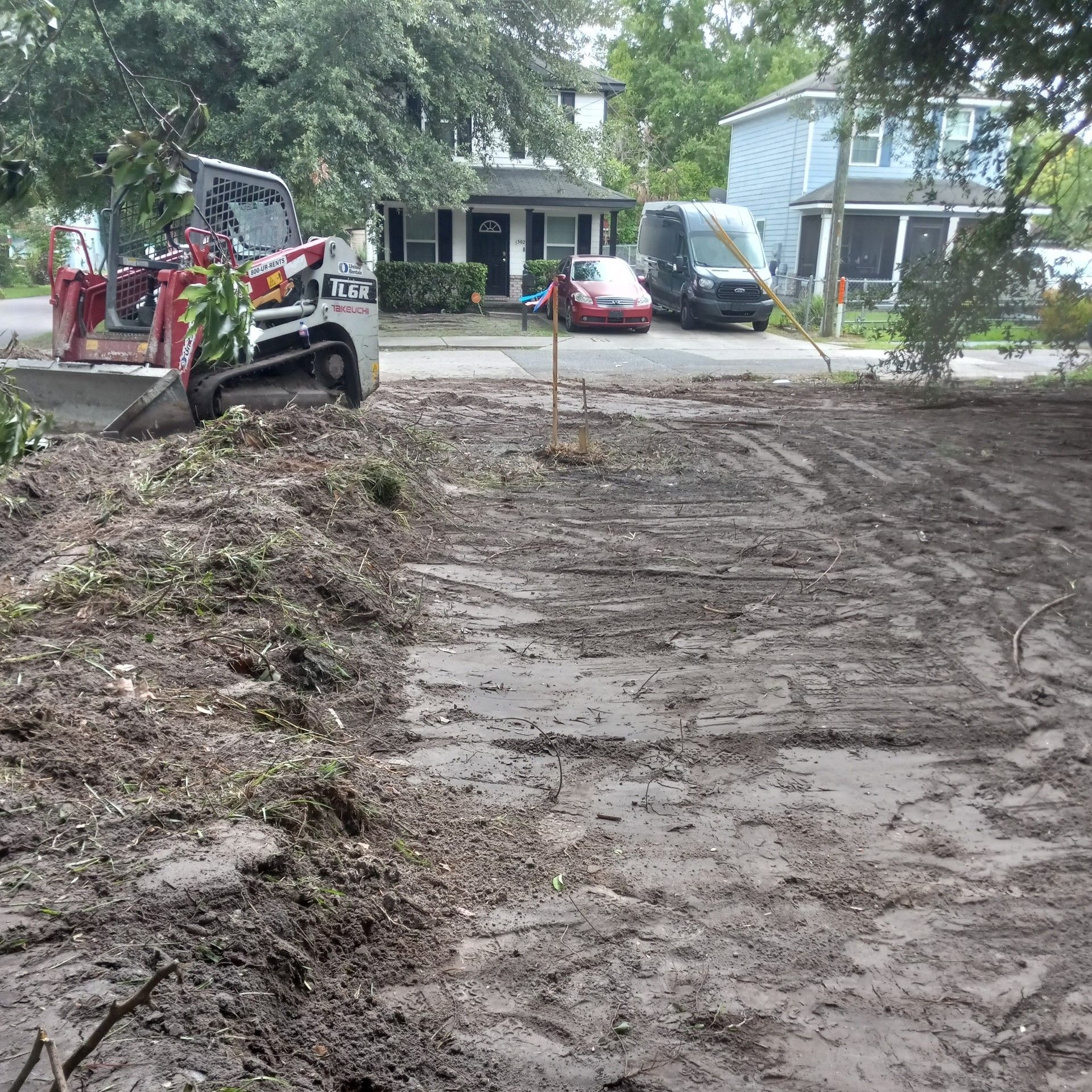 Construction site with a small excavator clearing muddy ground in front of parked cars and houses