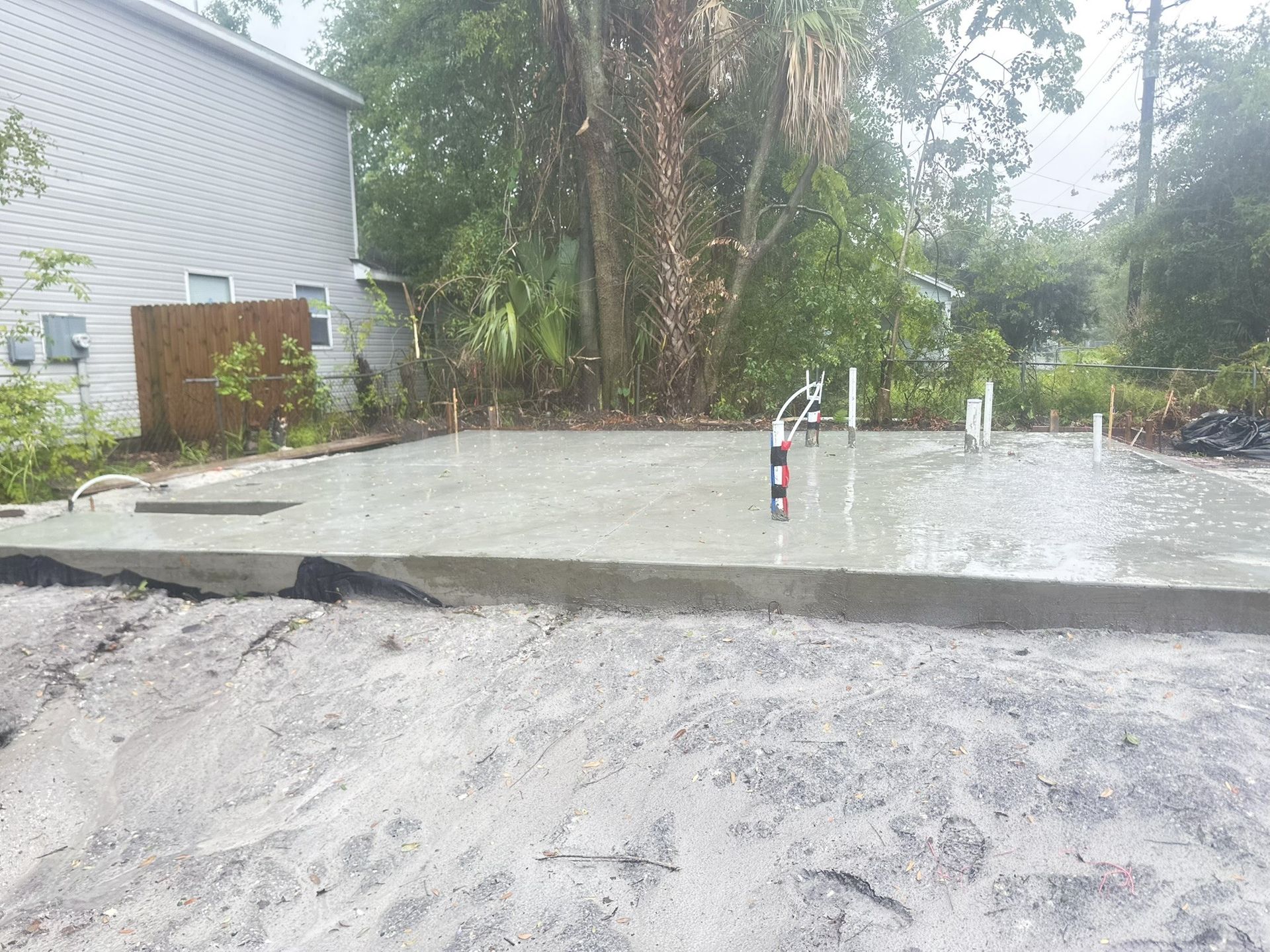 A freshly poured concrete foundation slab sits in a residential backyard next to a house, with exposed plumbing pipes.