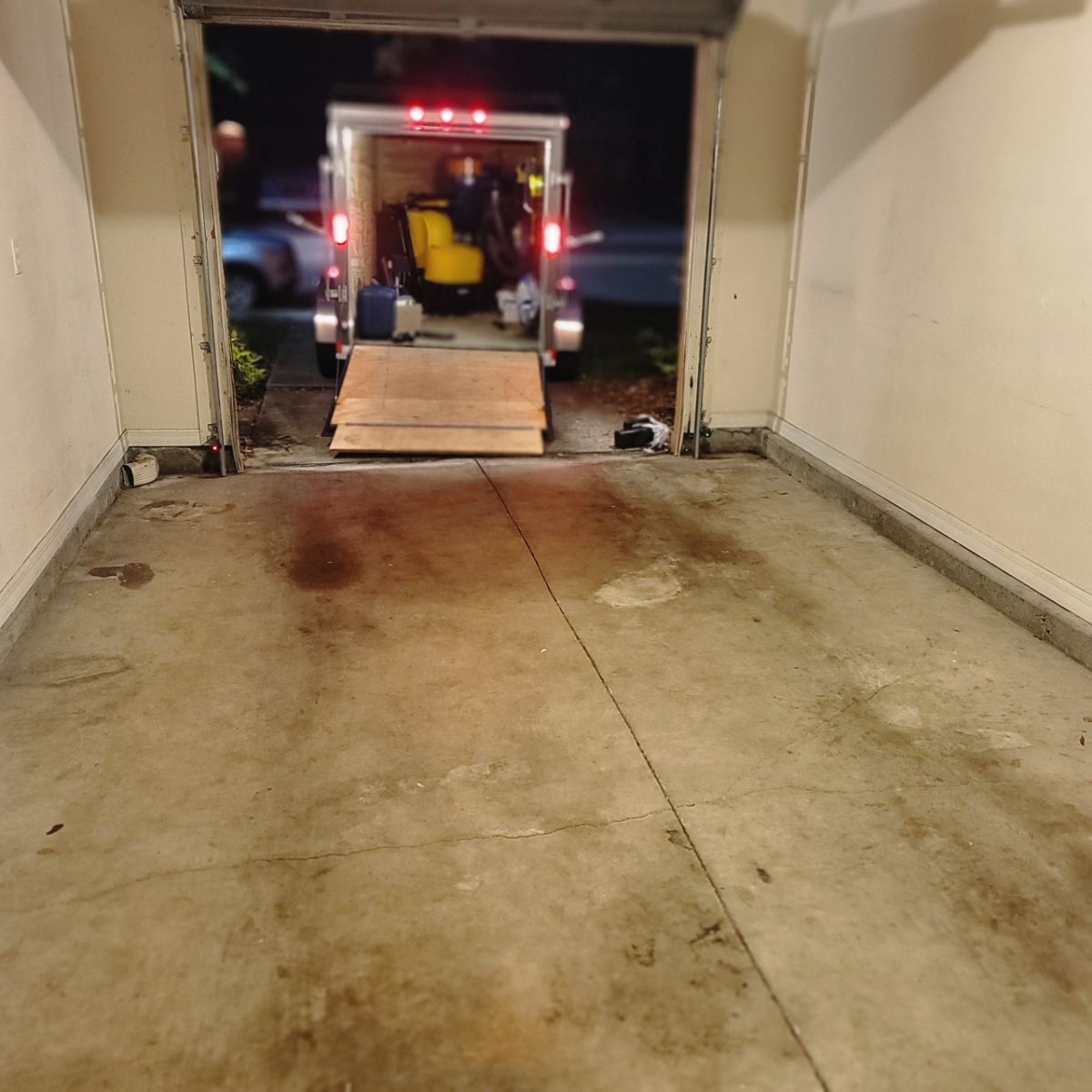 Empty garage with an open door; an epoxy trailer with a yellow machine and ramp is parked outside at night.