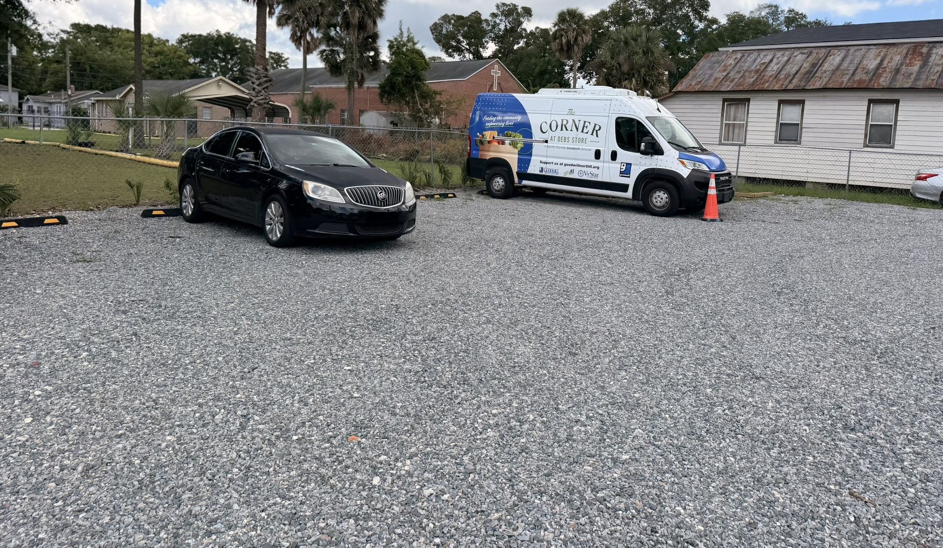 Black car and white service van parked on a gravel lot near houses, with orange cones nearby