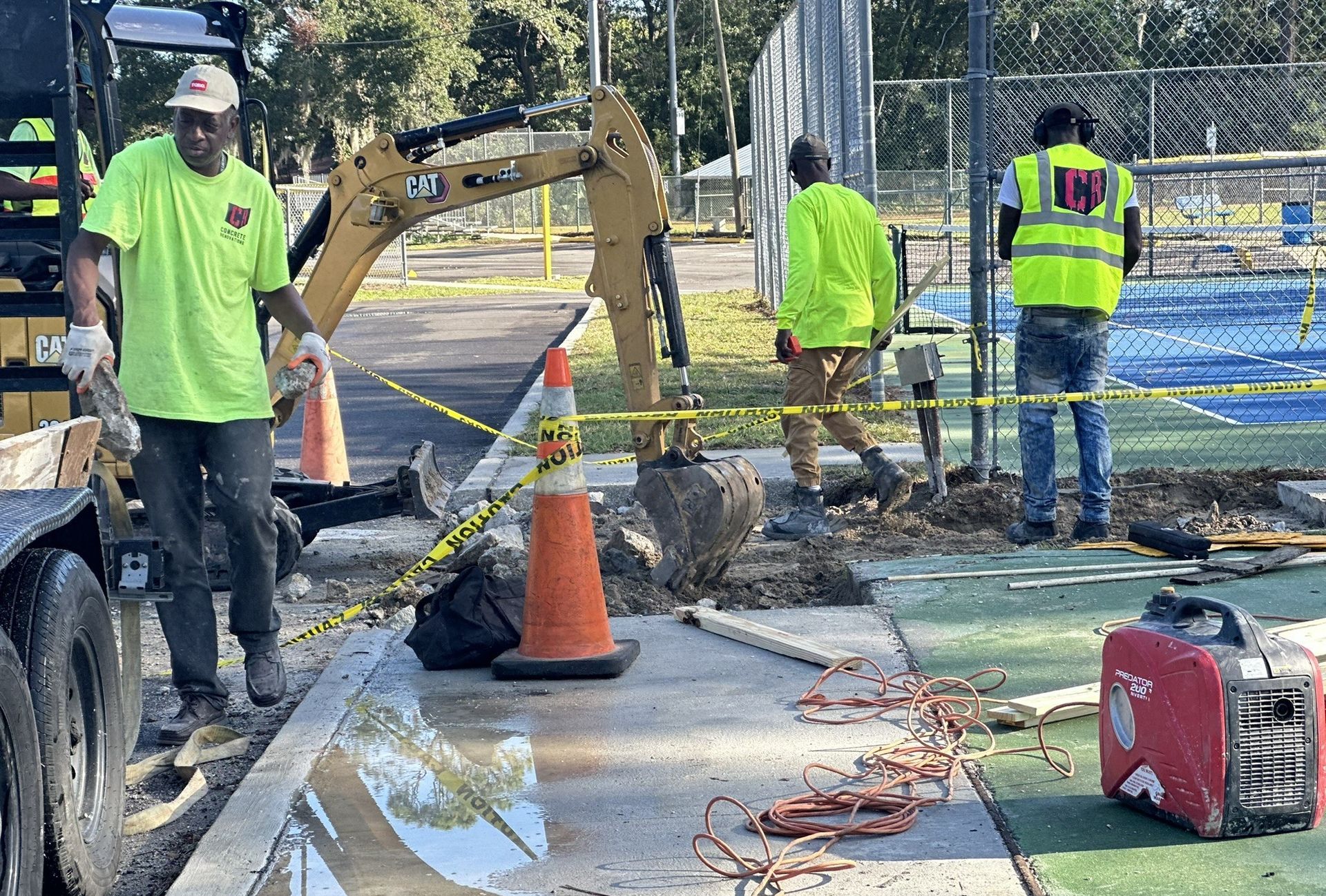 Roadwork crew repairing a sidewalk trench with a backhoe, orange cone, and tools on a sunny street.