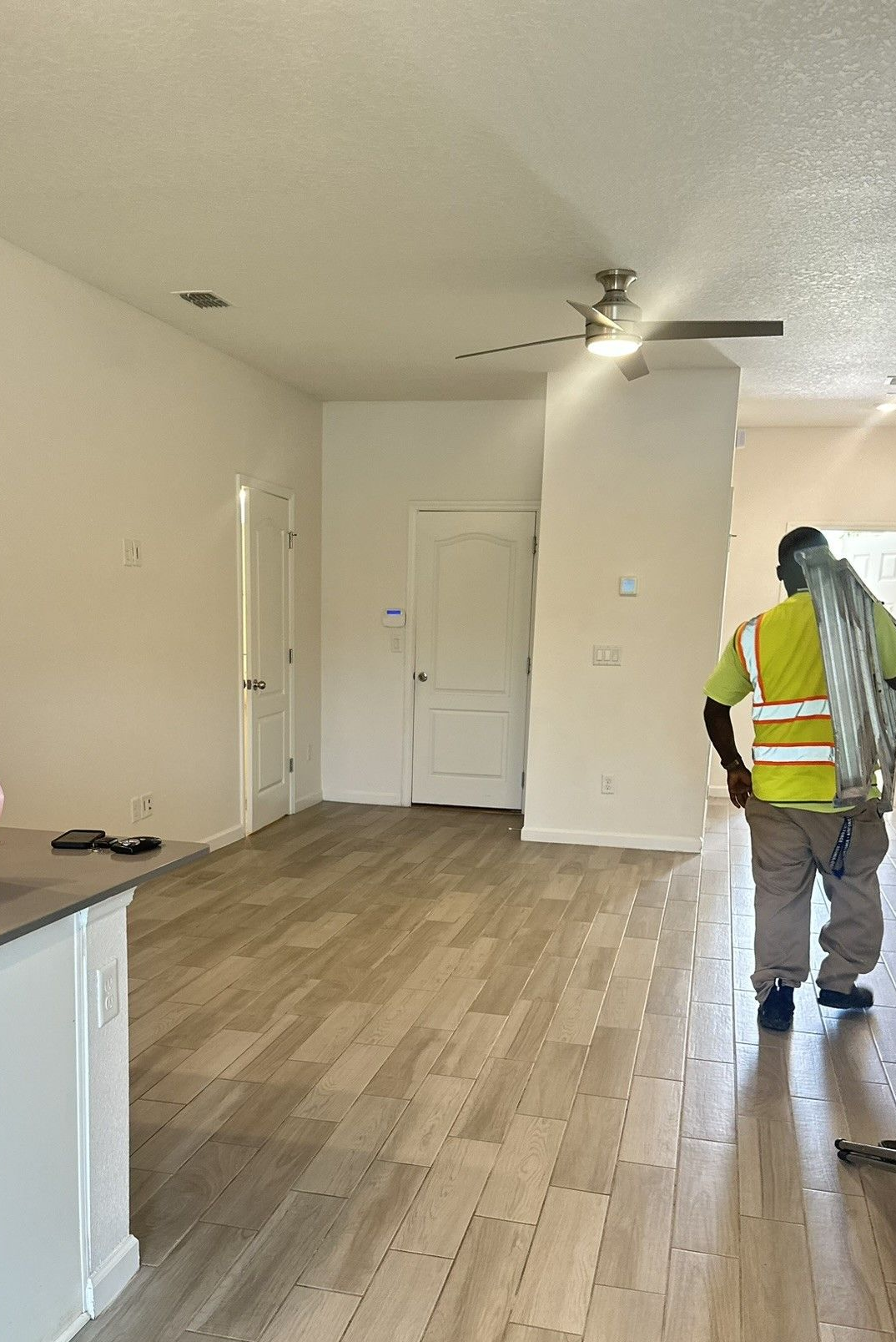 Worker carrying metal tubes in a freshly painted room with wood-look flooring and white walls, door, and a ceiling fan.