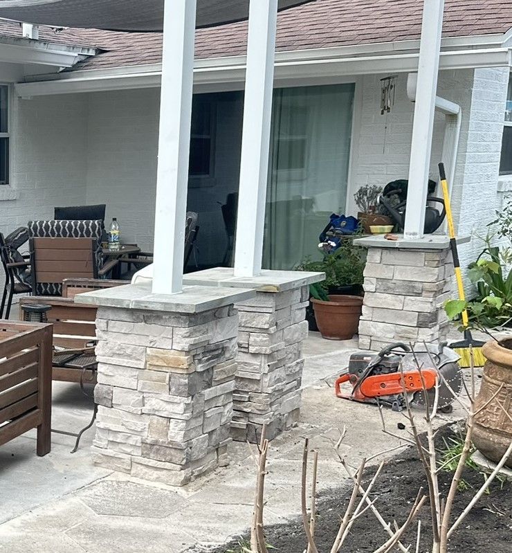 A patio area with white support columns on stone bases, outdoor furniture, a concrete saw, and potted plants near a house.