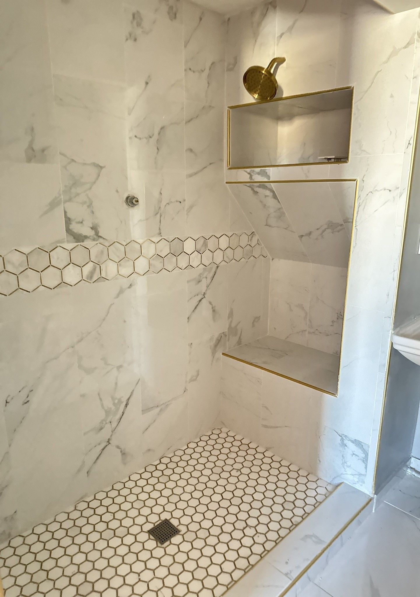 Marble-tiled shower installation with hexagon floor, built-in corner shelves, and a gold showerhead slot.