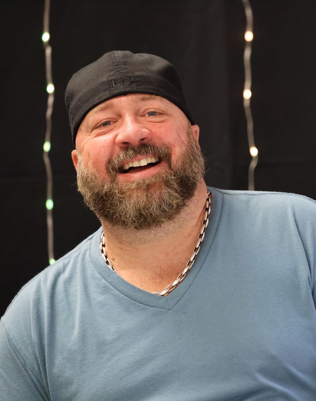 Spence Young smiling with a beard and black cap wearing a blue v-neck shirt and silver chain against a dark background.