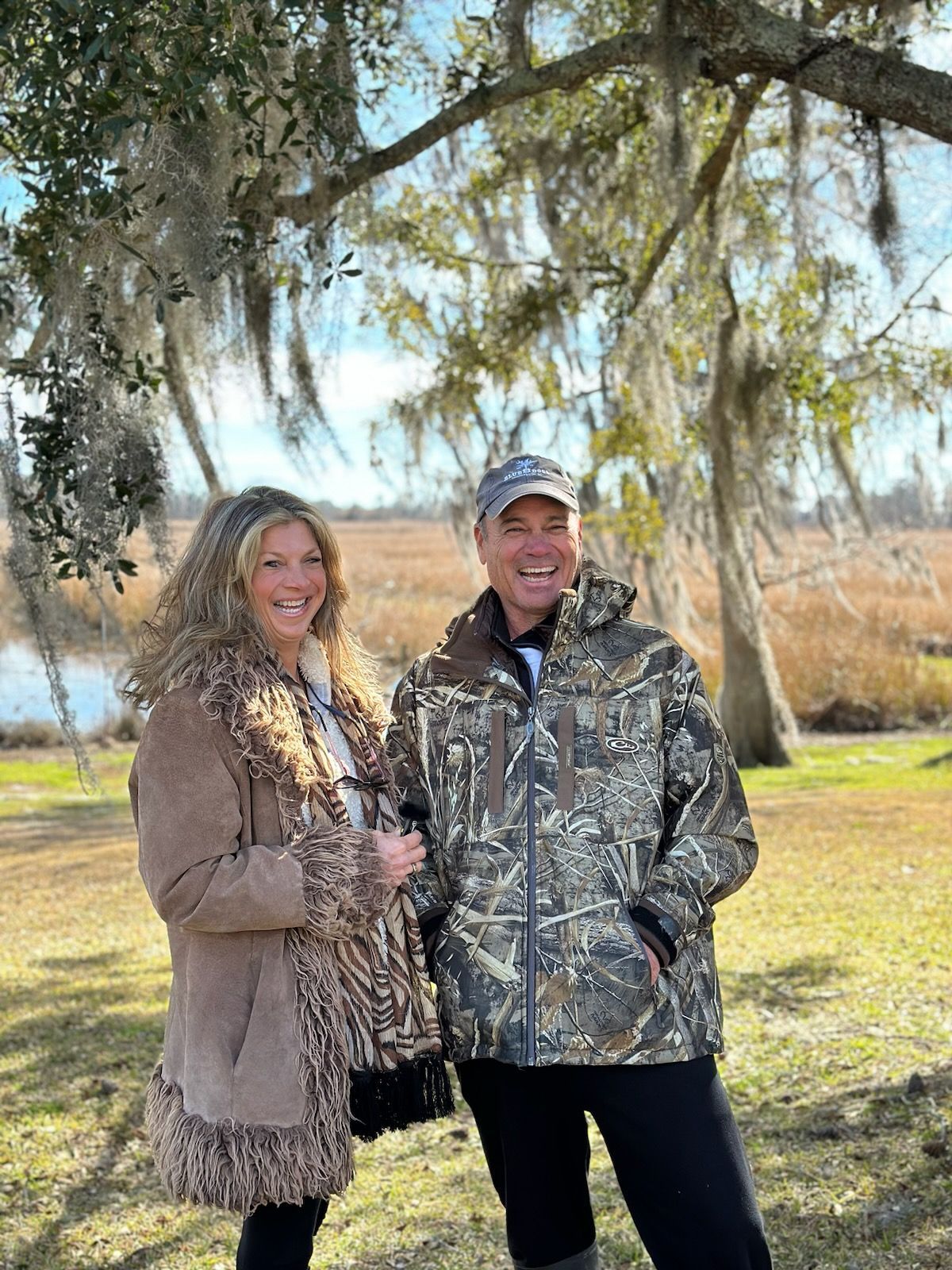 A smiling couple standing outdoors in a grassy area with moss-covered trees and water in the background.