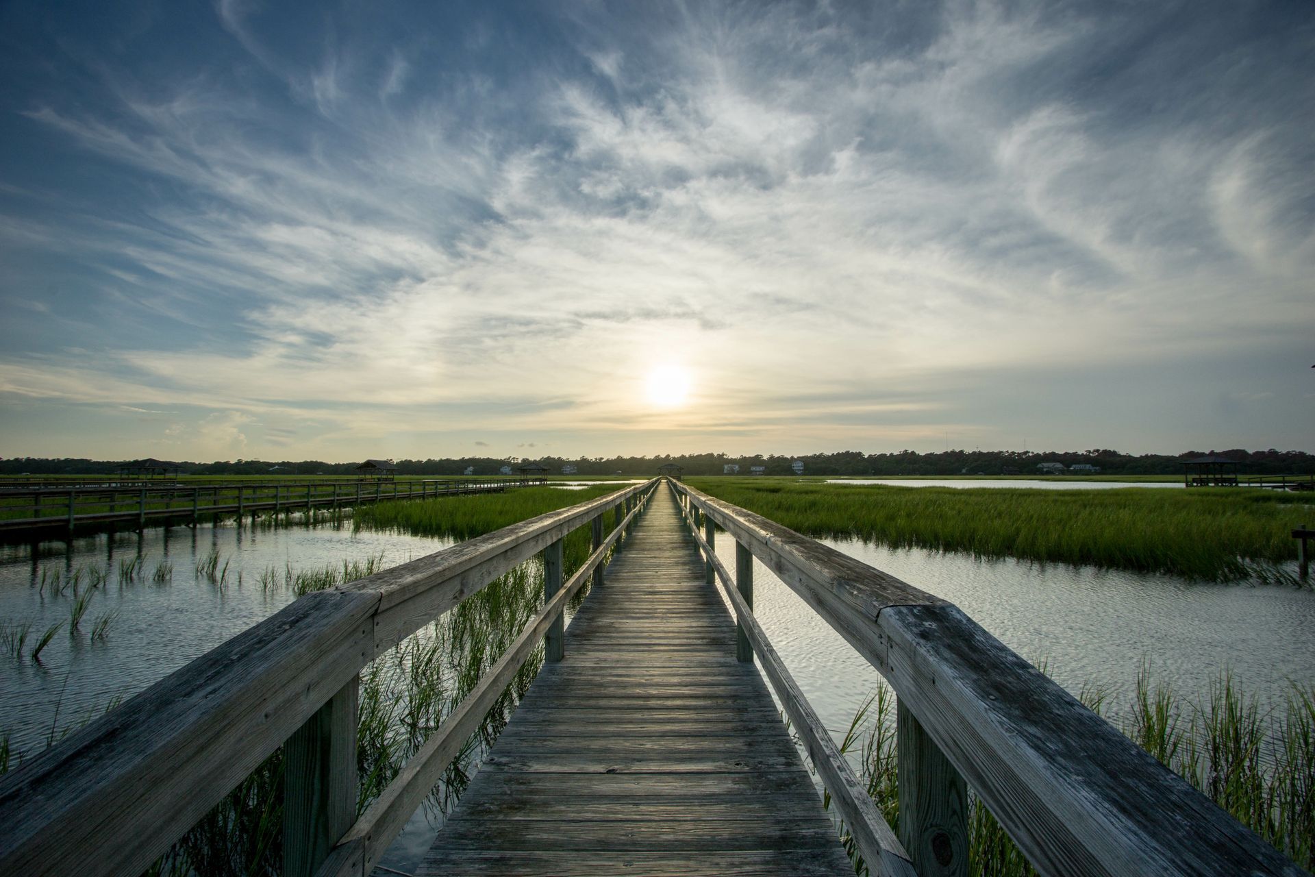 A wooden boardwalk stretches through a sunlit marsh under a vast, cloud-streaked sky at sunset.