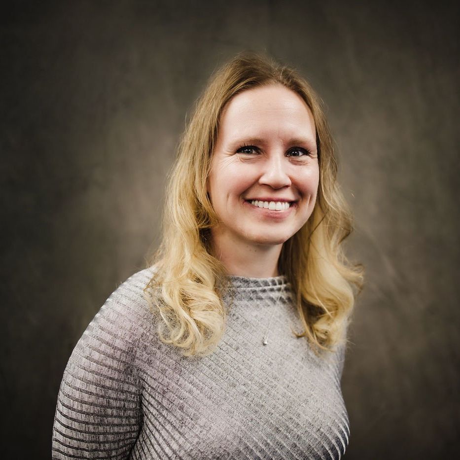 Smiling woman with blonde hair in a gray sweater against a dark studio background