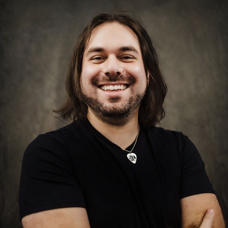 Smiling person with long dark hair wearing a black shirt and necklace against a gray backdrop