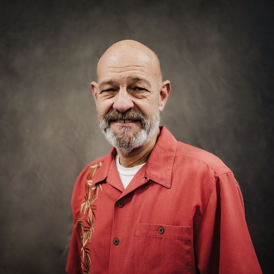 Smiling bald man with gray beard in red shirt against a dark studio backdrop