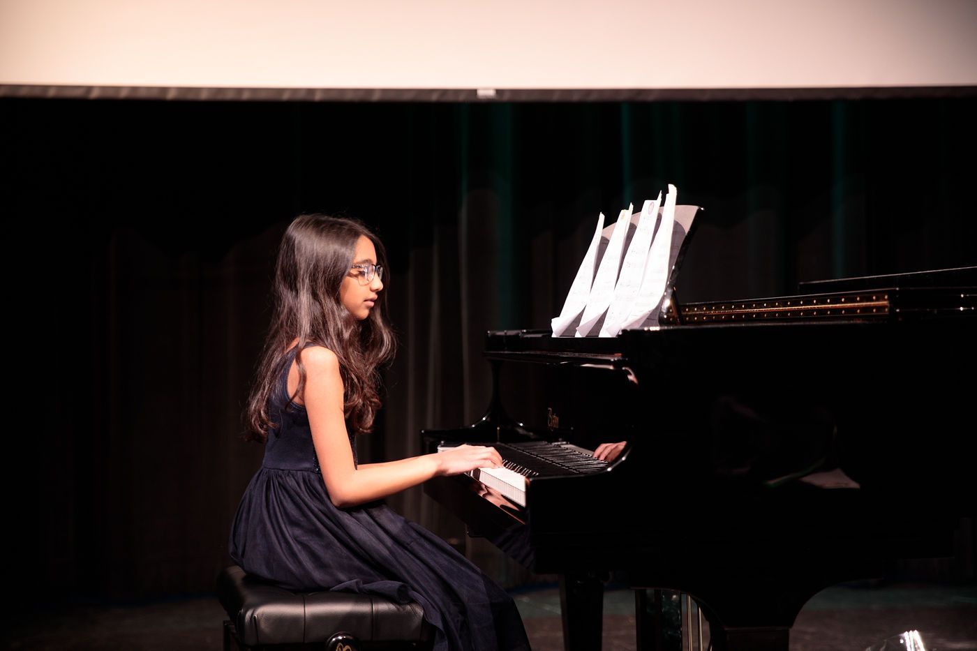 Young pianist in a dark dress playing a grand piano onstage with sheet music