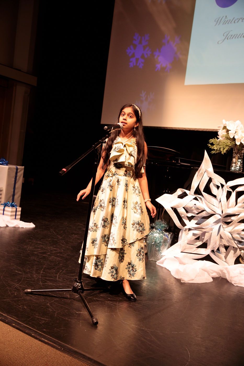 Singer performing on stage in a floral dress beside a microphone, with snowflake decorations and a piano backdrop.