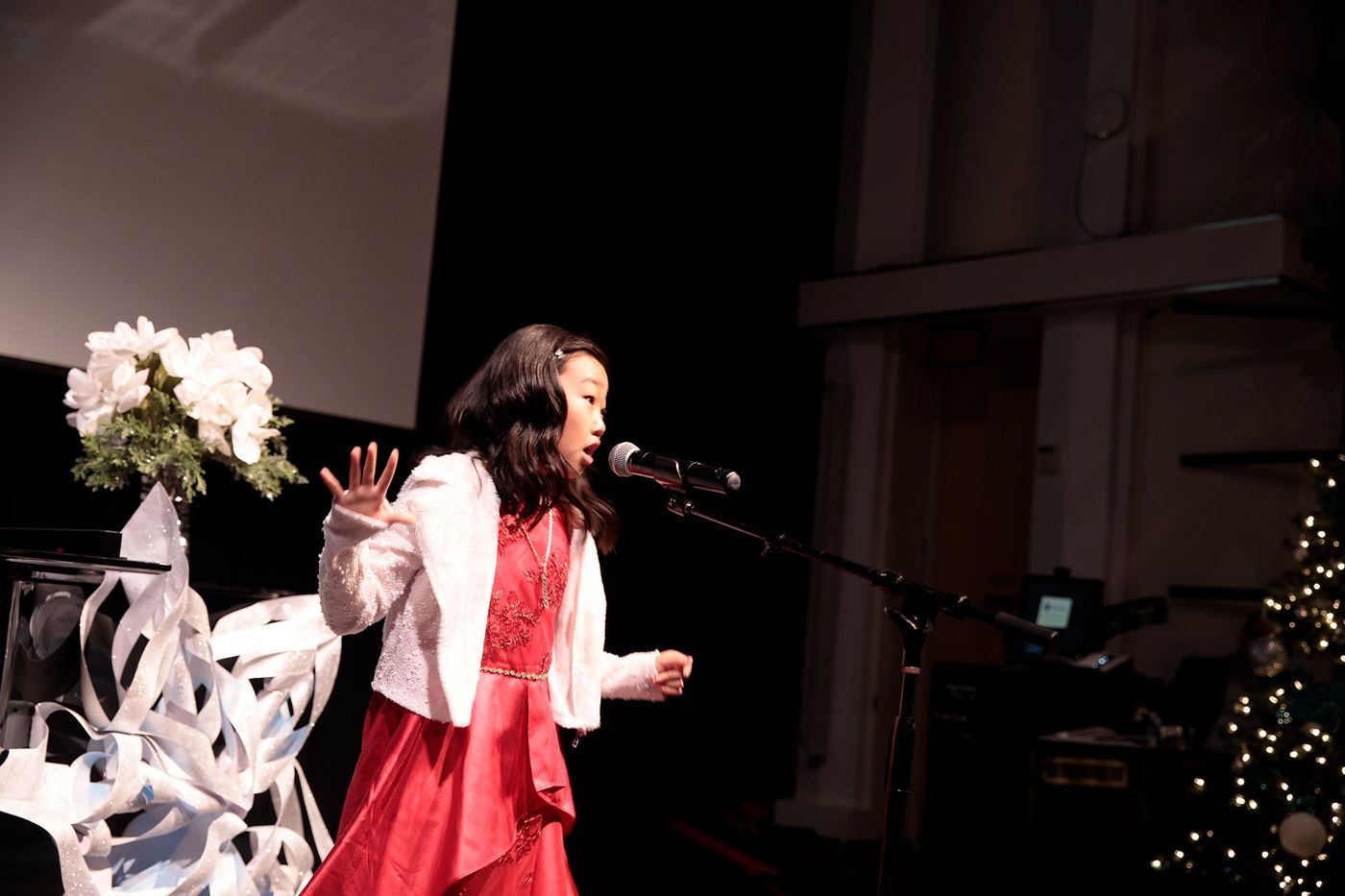 Singer performing on stage in a red dress and white jacket, speaking into a microphone with flowers nearby