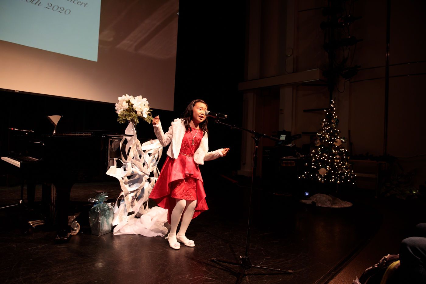 Child in red dress performing on a dimly lit stage beside a piano and Christmas tree