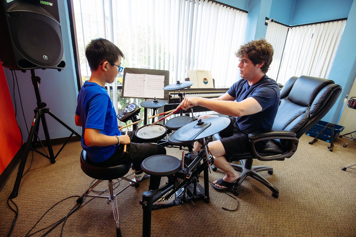 Two people playing electronic drums in a music room, with speakers and a laptop nearby.