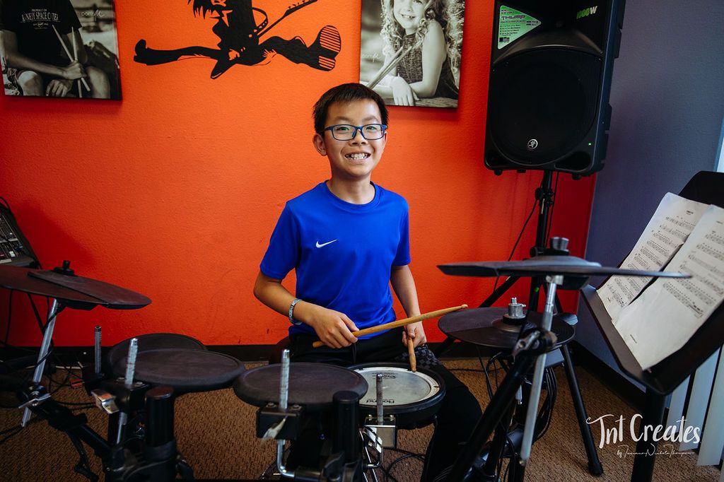 Young drummer in a blue shirt playing an electronic drum kit in a red-walled music room.