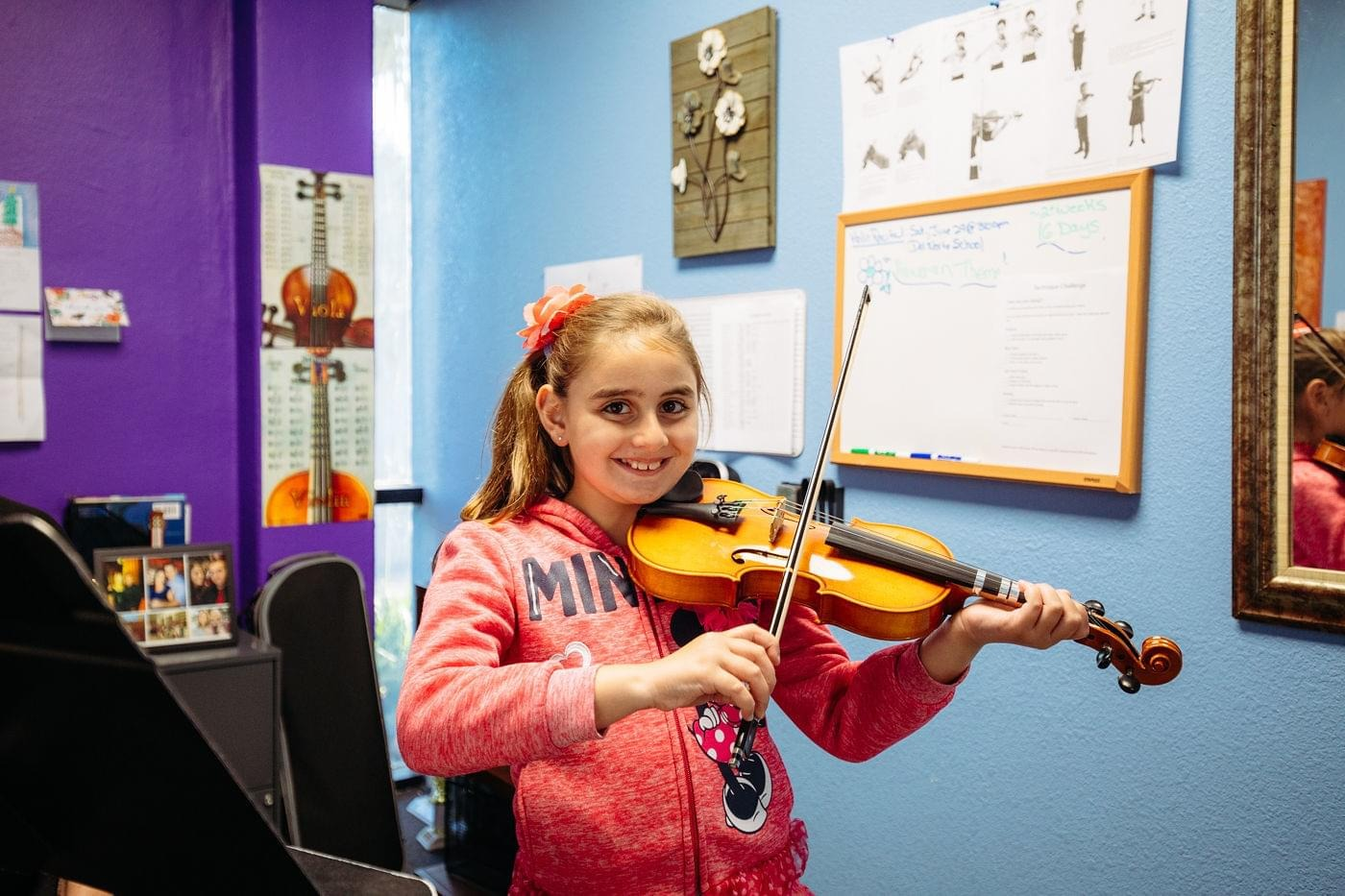 Child playing violin in a colorful music classroom, smiling while holding the instrument under her chin.