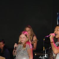 Children singing on stage with microphones, one in a silver top and pink headband.