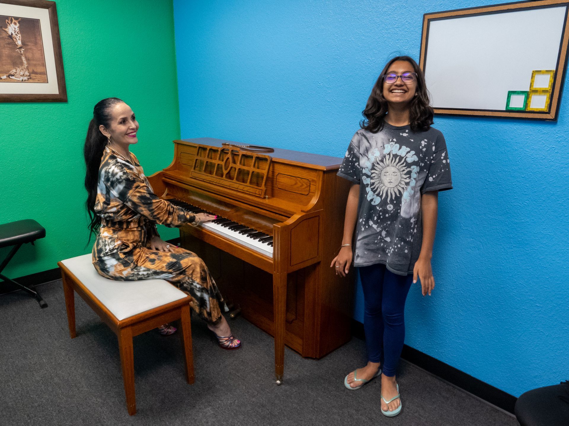 Two people in a colorful room, one seated at an upright piano and one standing beside a blue wall.