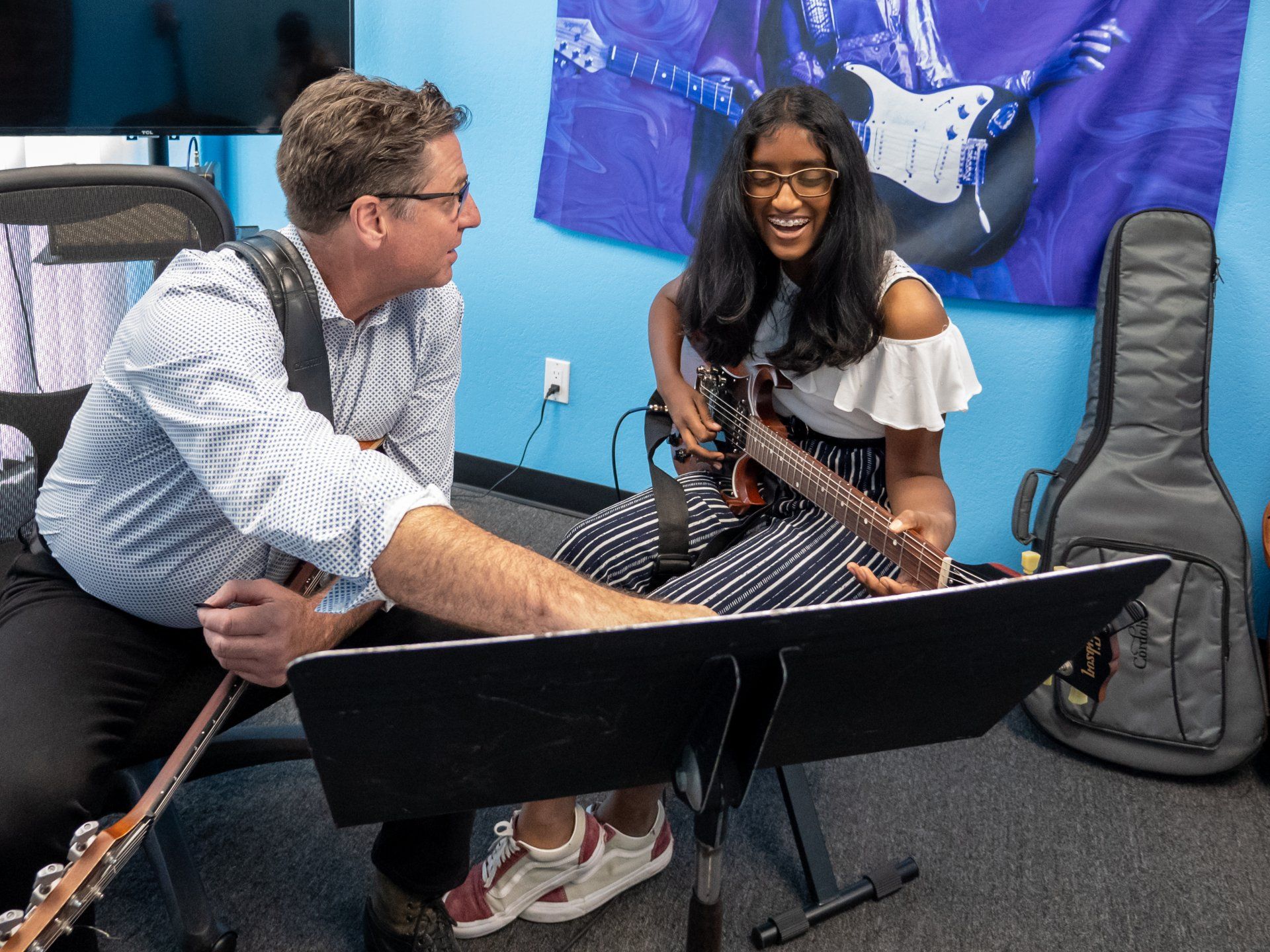 Two musicians rehearse with string instruments and music stands in a blue-walled room.