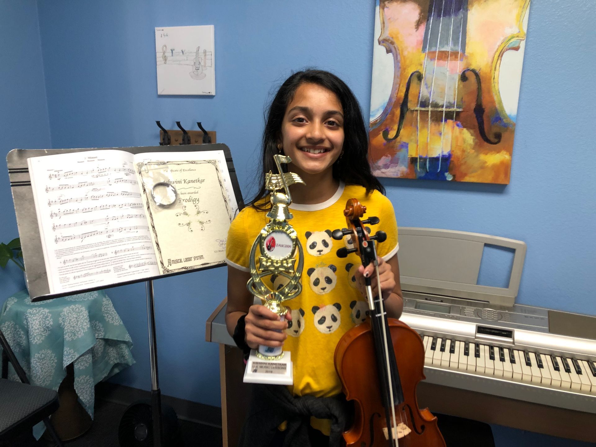 Smiling child holding a trophy and violin in a music room with sheet music and a keyboard.