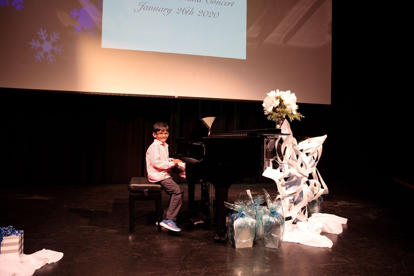 Child playing piano on stage beside a white floral display and decorative gift wrap