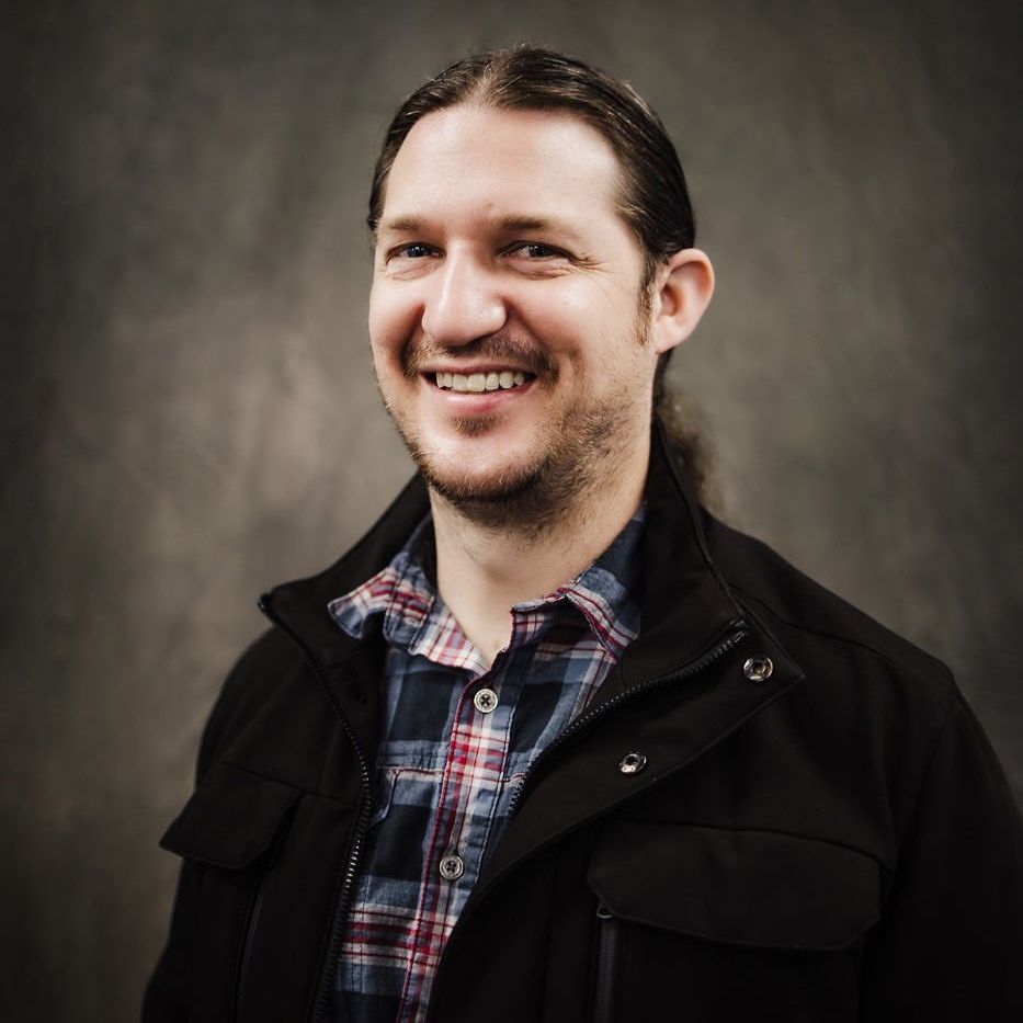 Smiling man in a plaid shirt and black jacket against a dark studio backdrop