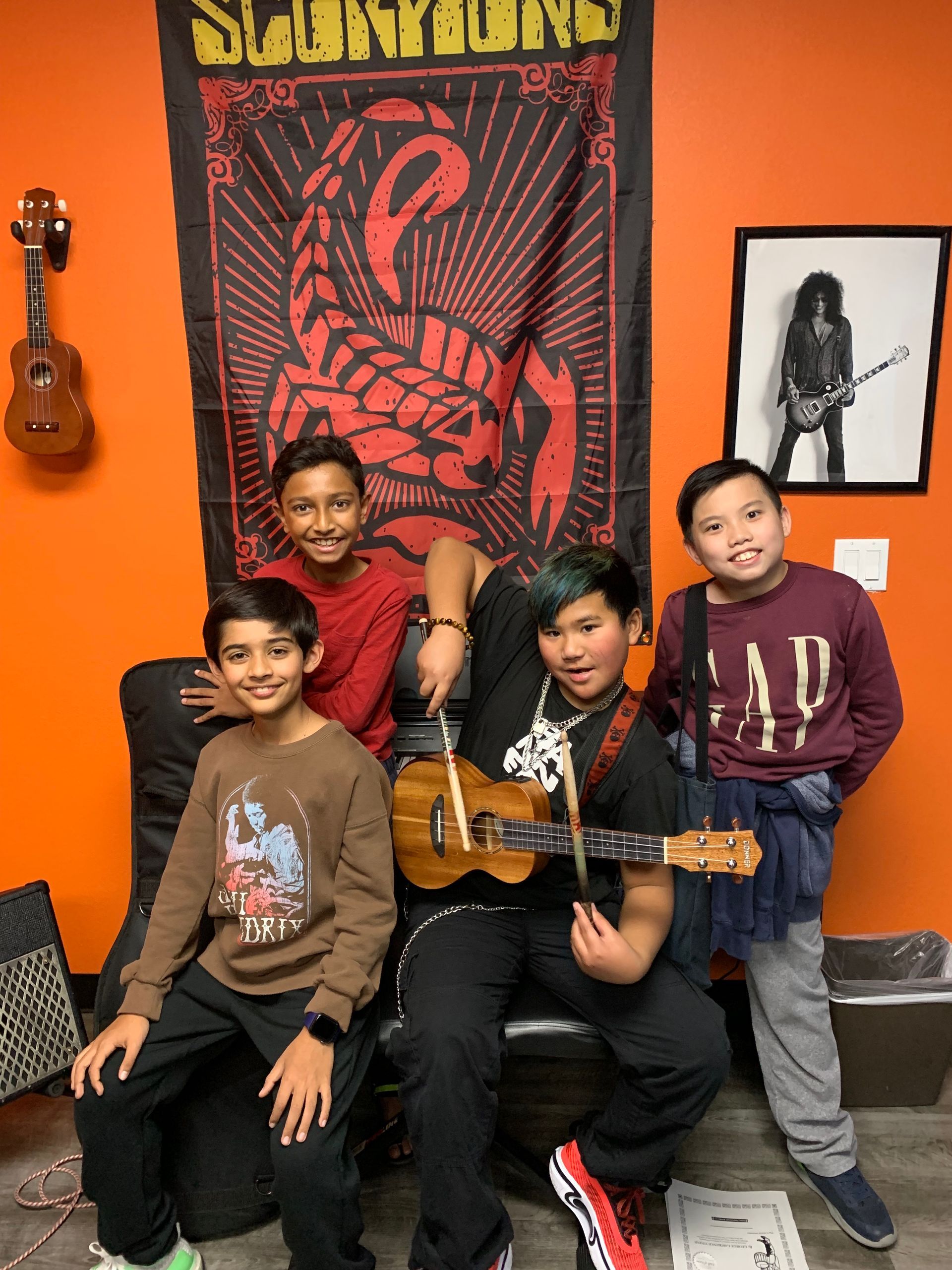 Four people posing with guitars in a bright orange music room, smiling in front of a band poster