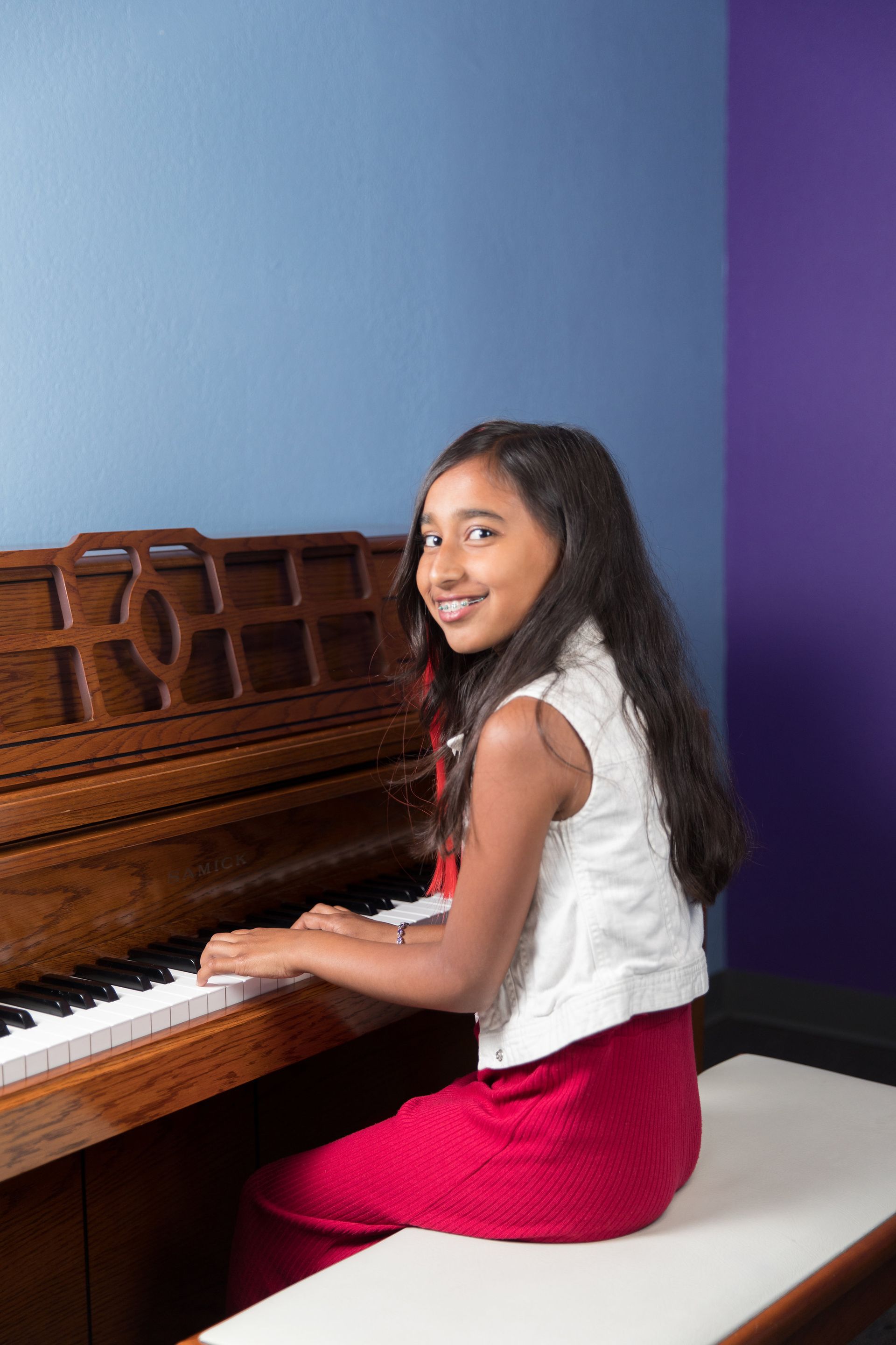 Girl seated at a piano bench, smiling and playing a wooden piano against a blue and purple wall.
