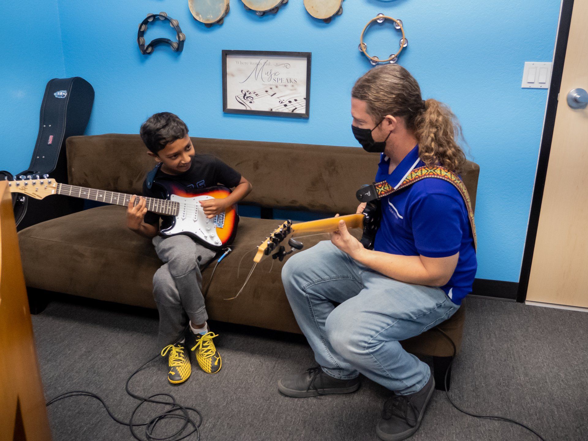 Two people seated on a couch play electric guitars in a blue-walled room with tambourines and a clock.