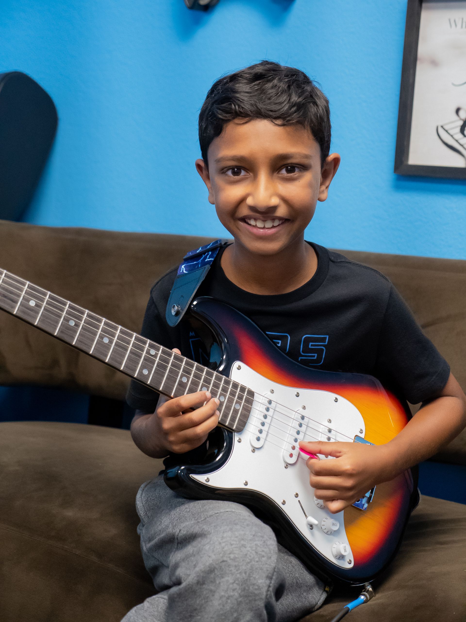 Child smiling while holding an electric guitar on a couch, with a blue wall behind.