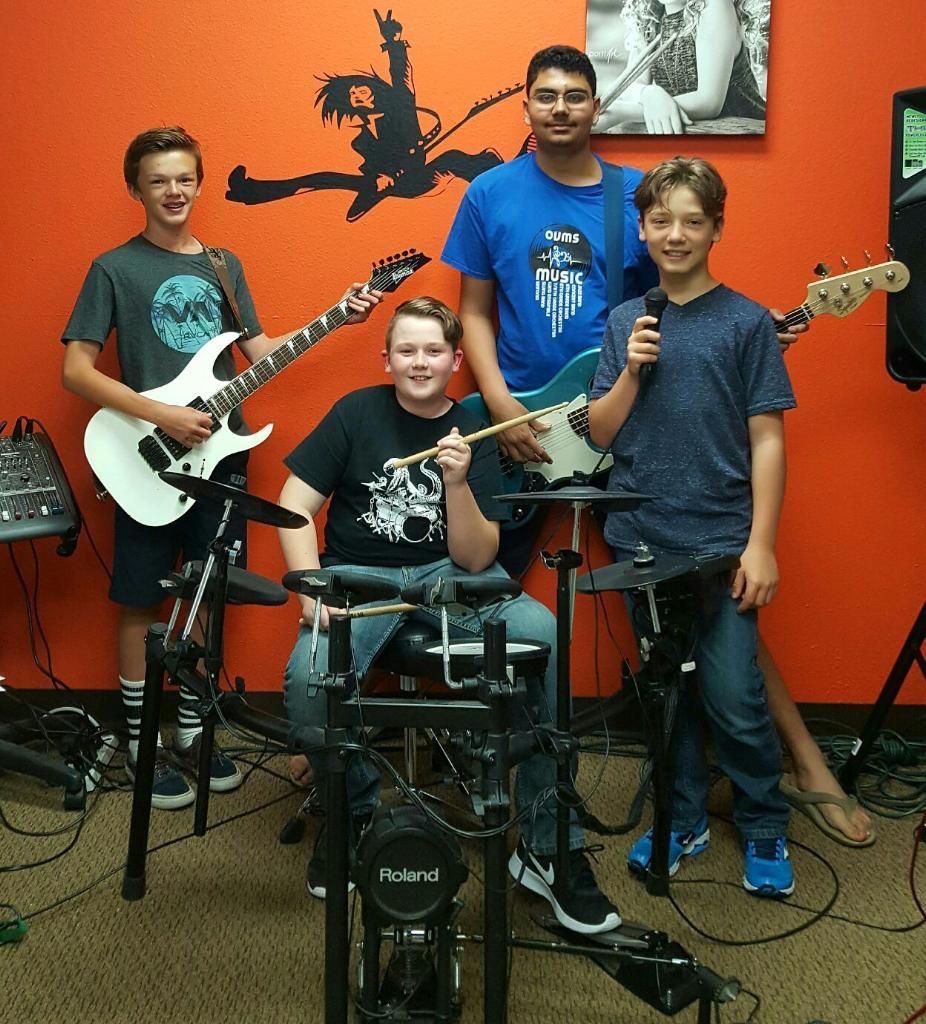Four young musicians posing with guitars and drum kit in a music room with orange walls
