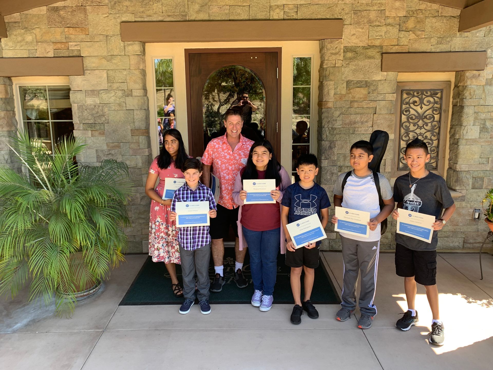 Seven children and one adult stand outside a stone building, holding certificates and smiling.