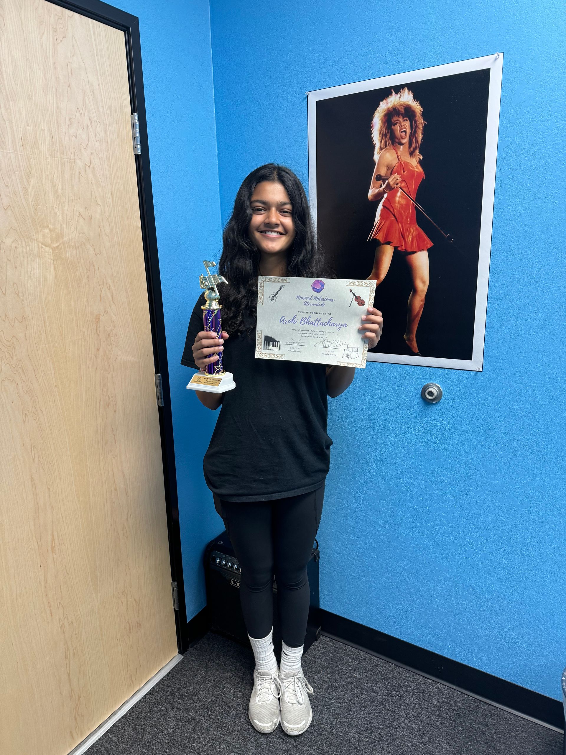 Person holding a trophy and certificate, smiling in a blue room with a poster on the wall