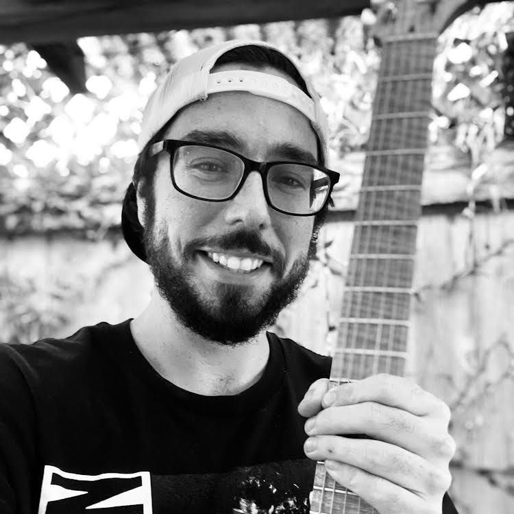 Smiling person in glasses and a backward cap holding a guitar outdoors