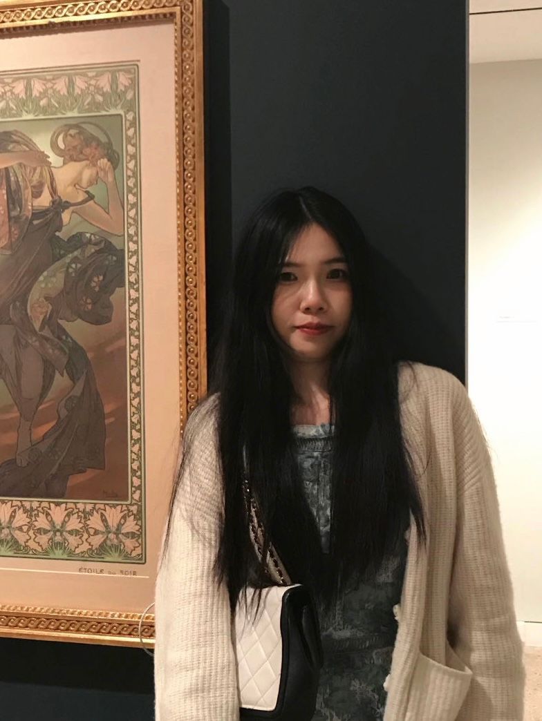 Woman standing indoors beside a framed painting, wearing a light cardigan and dark top.