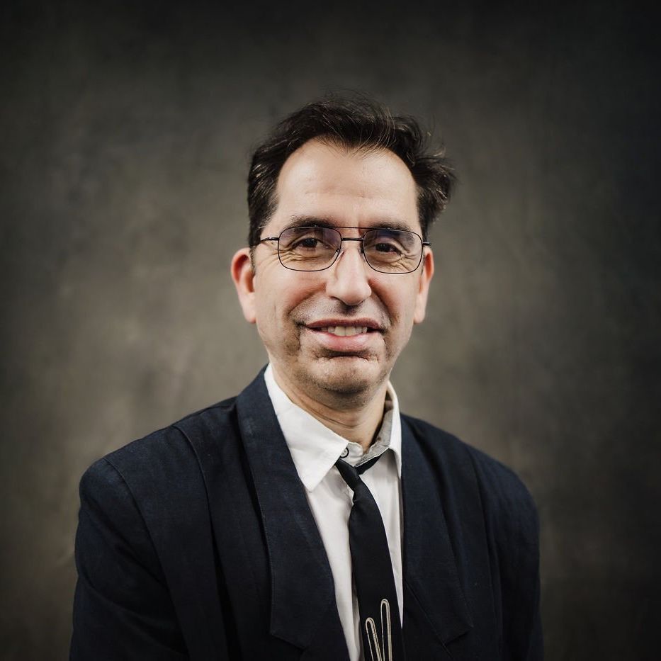 Portrait of a smiling person in a dark jacket, white shirt, and tie against a gray studio backdrop