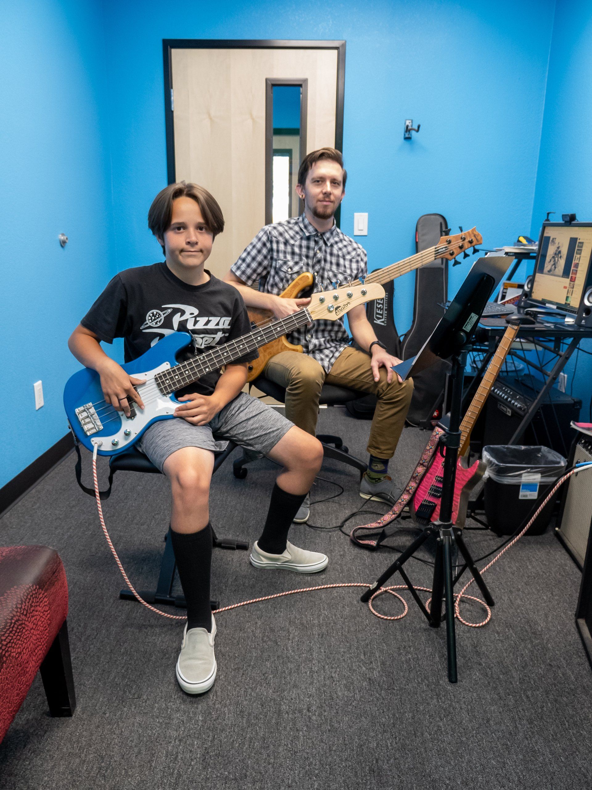 Two musicians sit in a studio holding electric guitars, with amps and music stands nearby.