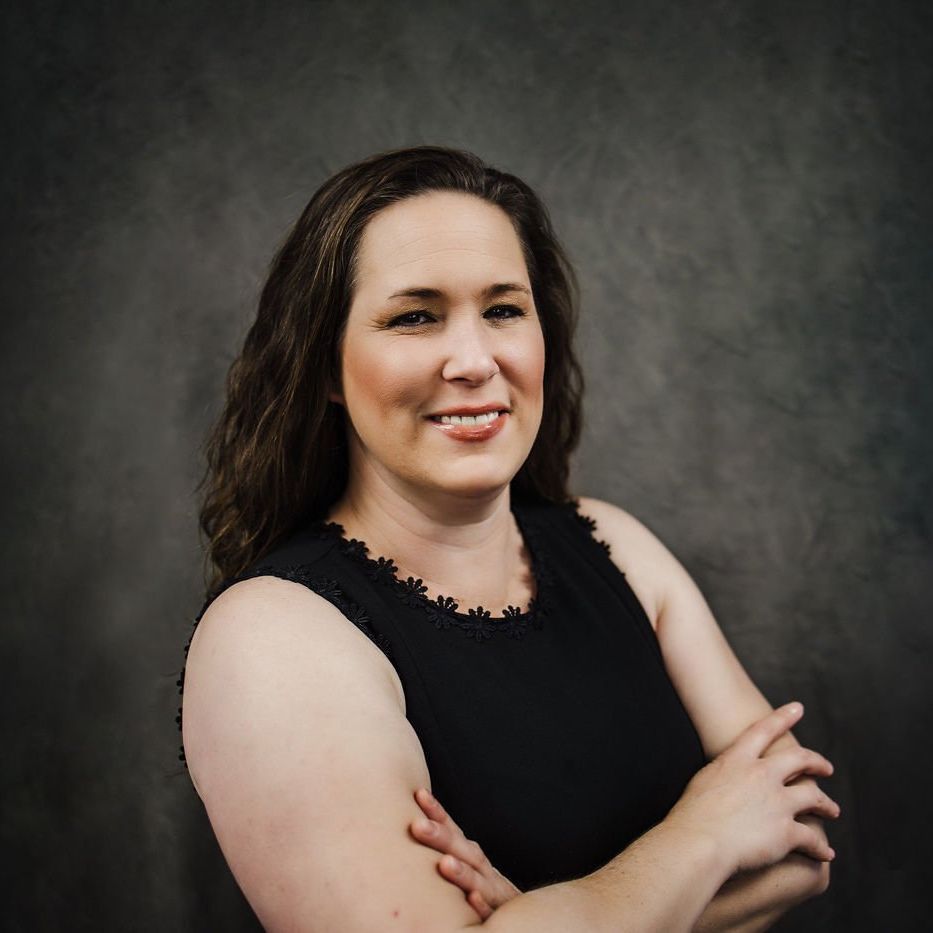 Portrait of a smiling woman in a black sleeveless top with arms crossed against a gray studio backdrop