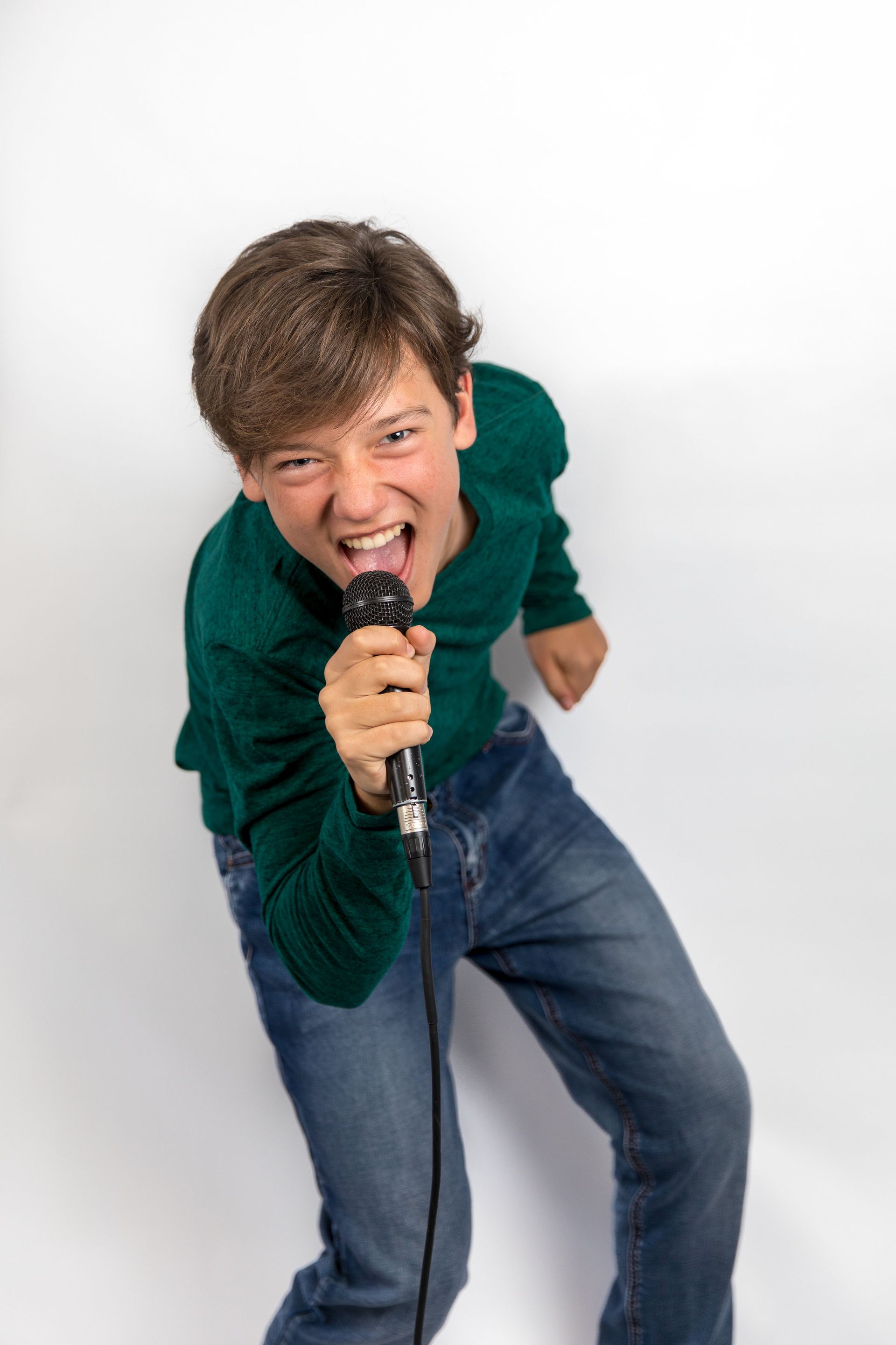 Person singing into a microphone, leaning forward in a green shirt and jeans against a white background.