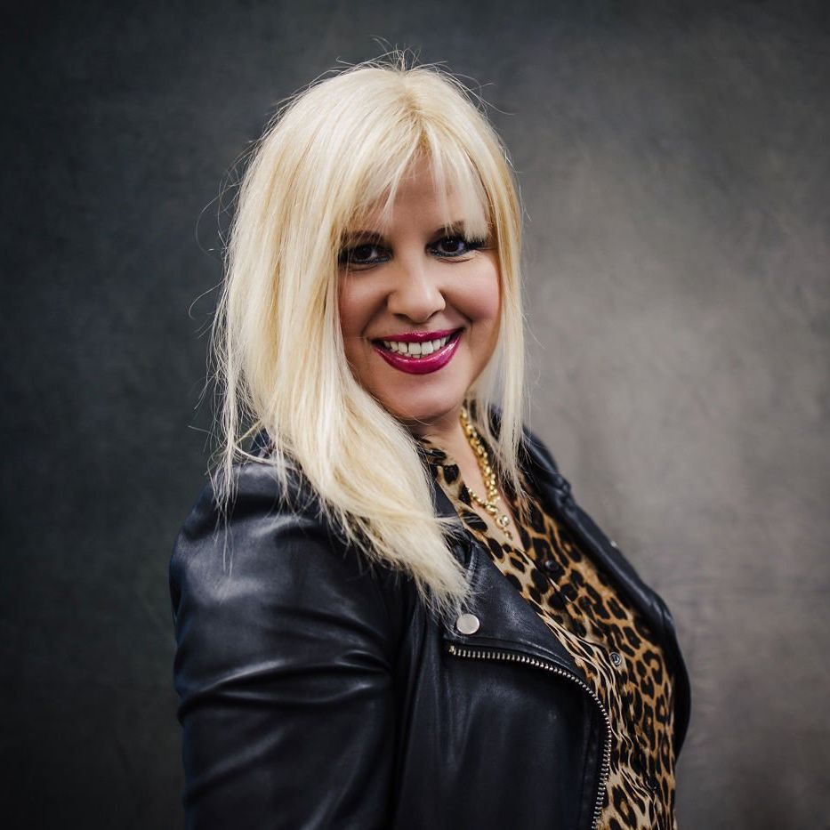 Blonde woman in a black leather jacket and leopard-print top smiling against a dark studio backdrop