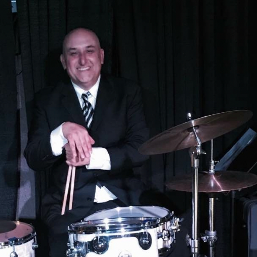 Smiling drummer in a suit, leaning on a snare drum beside cymbals on a dark stage.