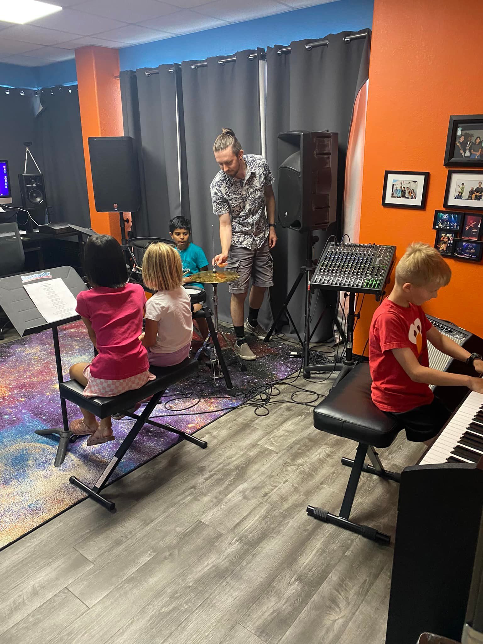 Music class with children seated at keyboards while a teacher stands near microphones on a small stage