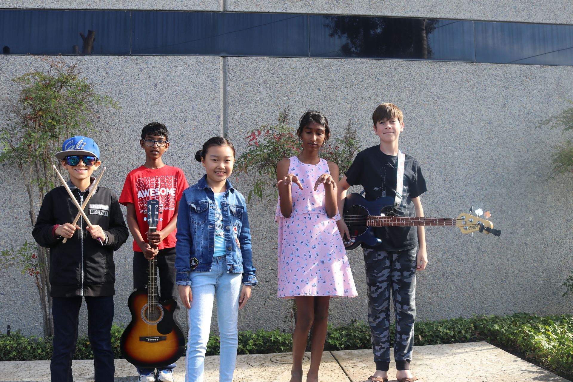 Five children stand outdoors with musical instruments, including guitars and a violin, in front of a wall.