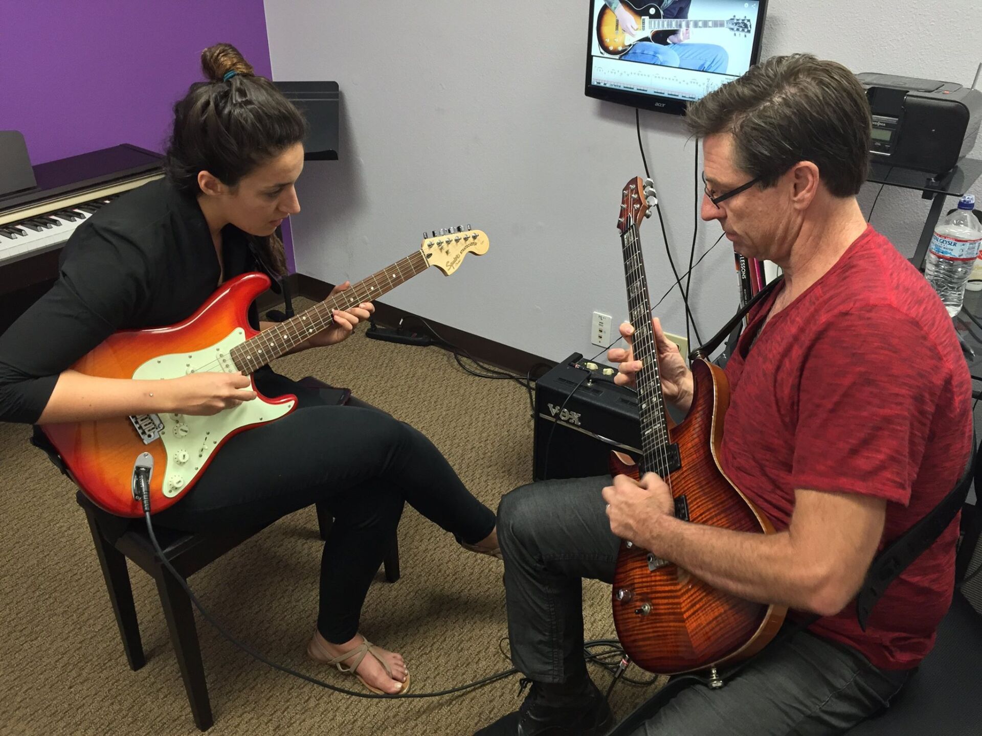 Two people seated, playing guitars in a room with a TV and purple wall.